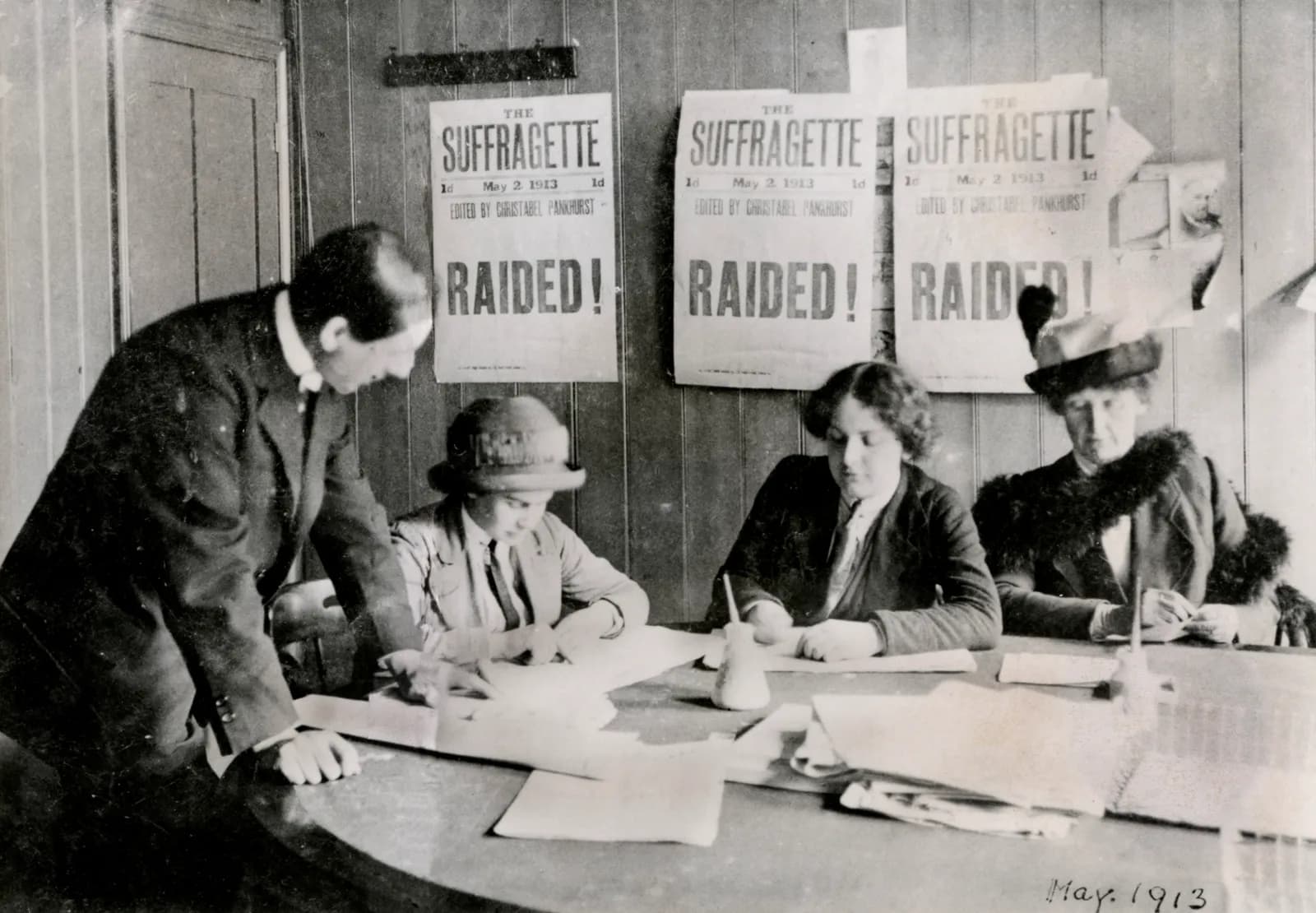 A group of suffragettes, including a man and three women, are gathered around a table in an office setting, with posters of 'The Suffragette' newspaper with large 'RAIDED!' text displayed on the wall behind them.