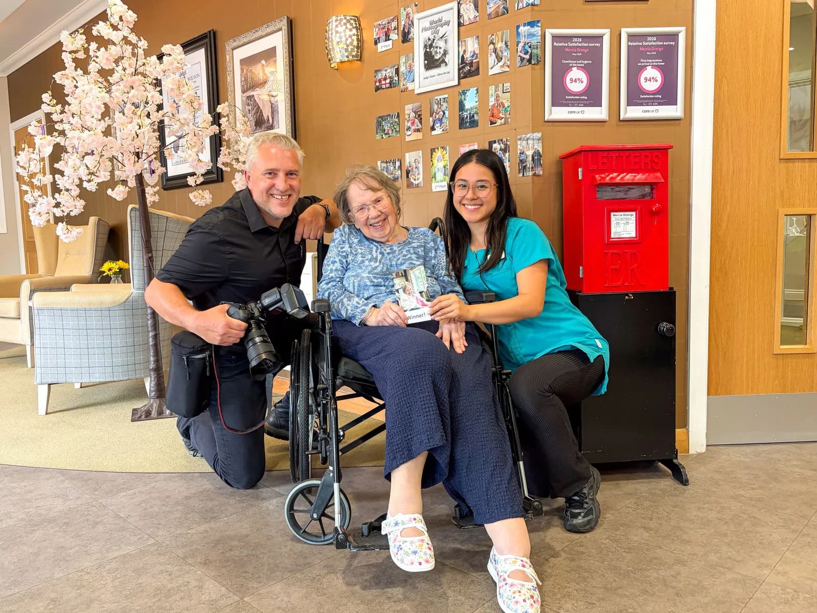 A man kneeling with a camera, an elderly woman in a wheelchair, and a younger woman in scrubs smiling together.