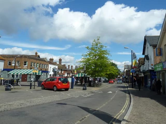 A quiet town square with a red car parked near green and white striped awnings and several buildings under a blue sky with fluffy white clouds.