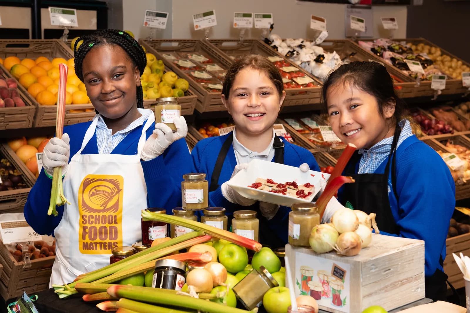 Three smiling students in aprons stand behind tables laden with produce and jars of food, holding up rhubarb, a jar, and a tray of small bites.