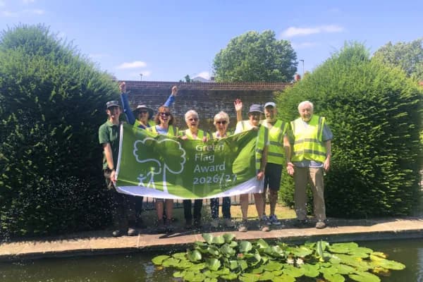 A group of cheerful people of various ages, some wearing high-visibility vests, hold up a green banner that reads "Green Flag Award 2026/27" in front of a pond with lily pads and green bushes on a sunny day.