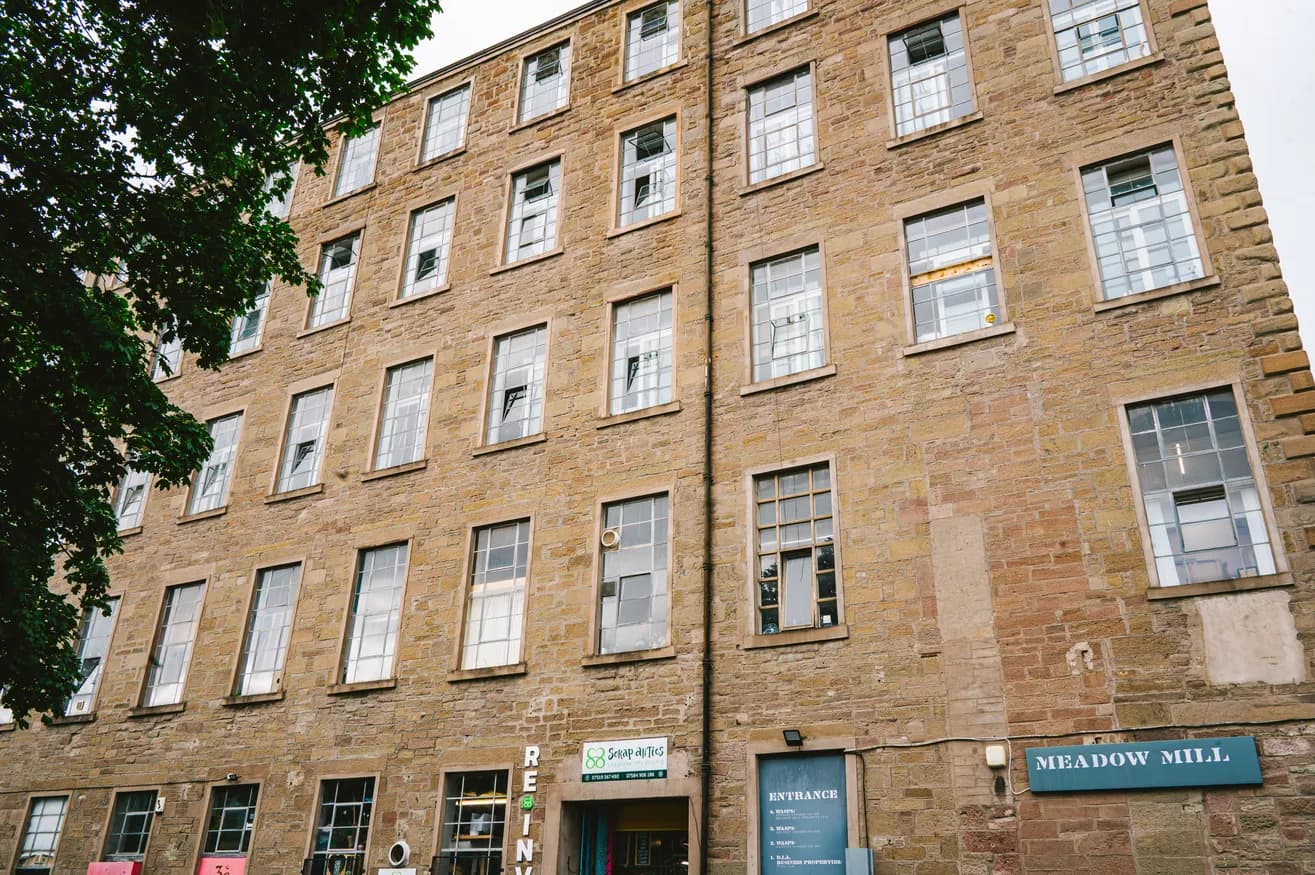 A tall, old brick building with many windows, some of which are open, with a sign for "Meadow Mill" near the bottom right.