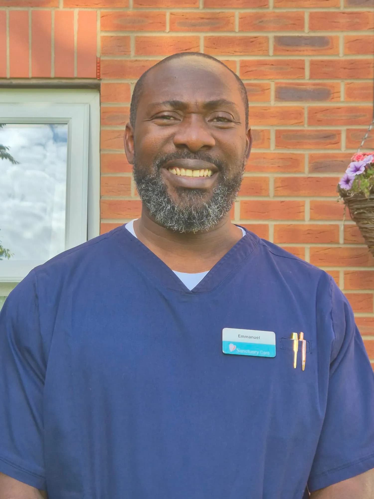 A man with a beard and a blue scrub top, smiling at the camera, with a brick wall behind him.