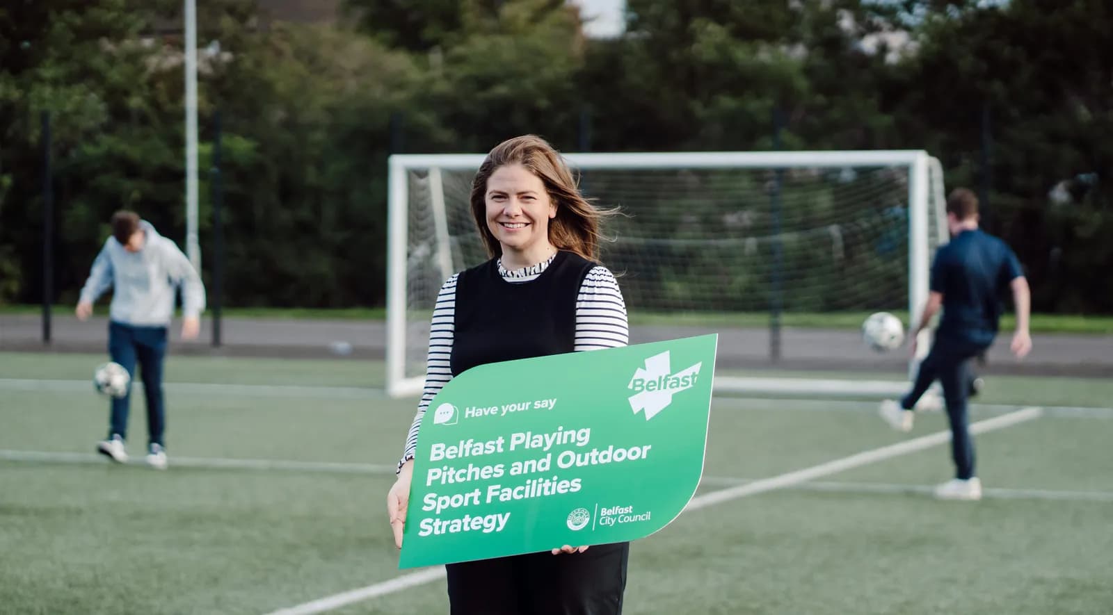 A smiling woman holding a green sign about Belfast playing pitches and outdoor sport facilities, standing on a football pitch with two men playing soccer in the background.