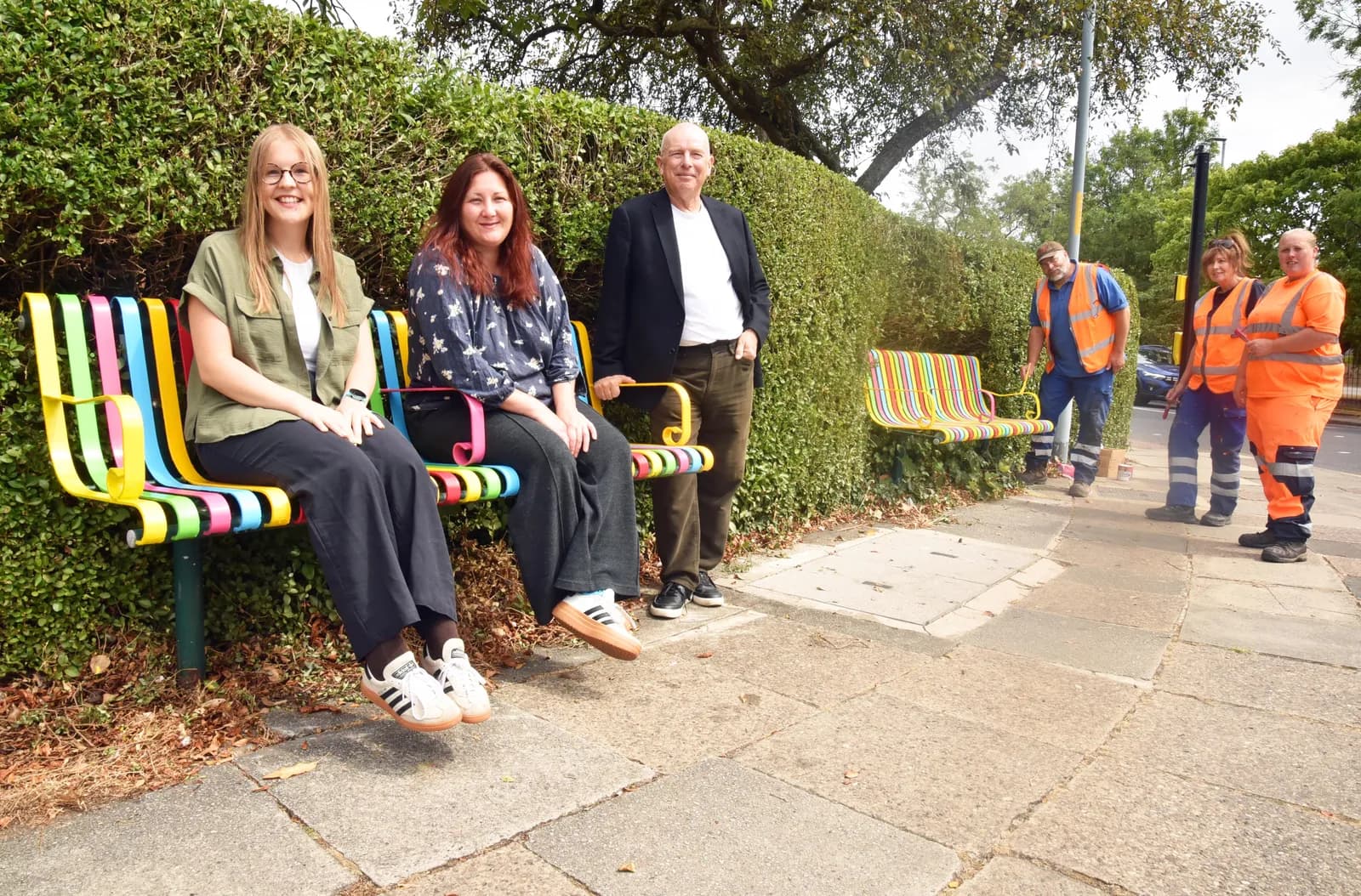 Two women and a man sit and stand by colorful benches in front of a green hedge, with three road workers in orange vests standing further down the path.