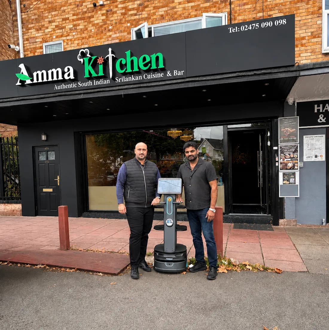 Two men stand in front of the Amma Kitchen restaurant with a gray service robot between them.