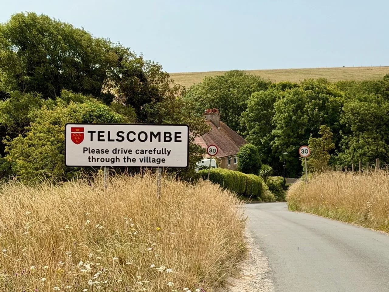 A "Telscombe - Please drive carefully through the village" sign stands beside a rural road, with a building and fields beyond.