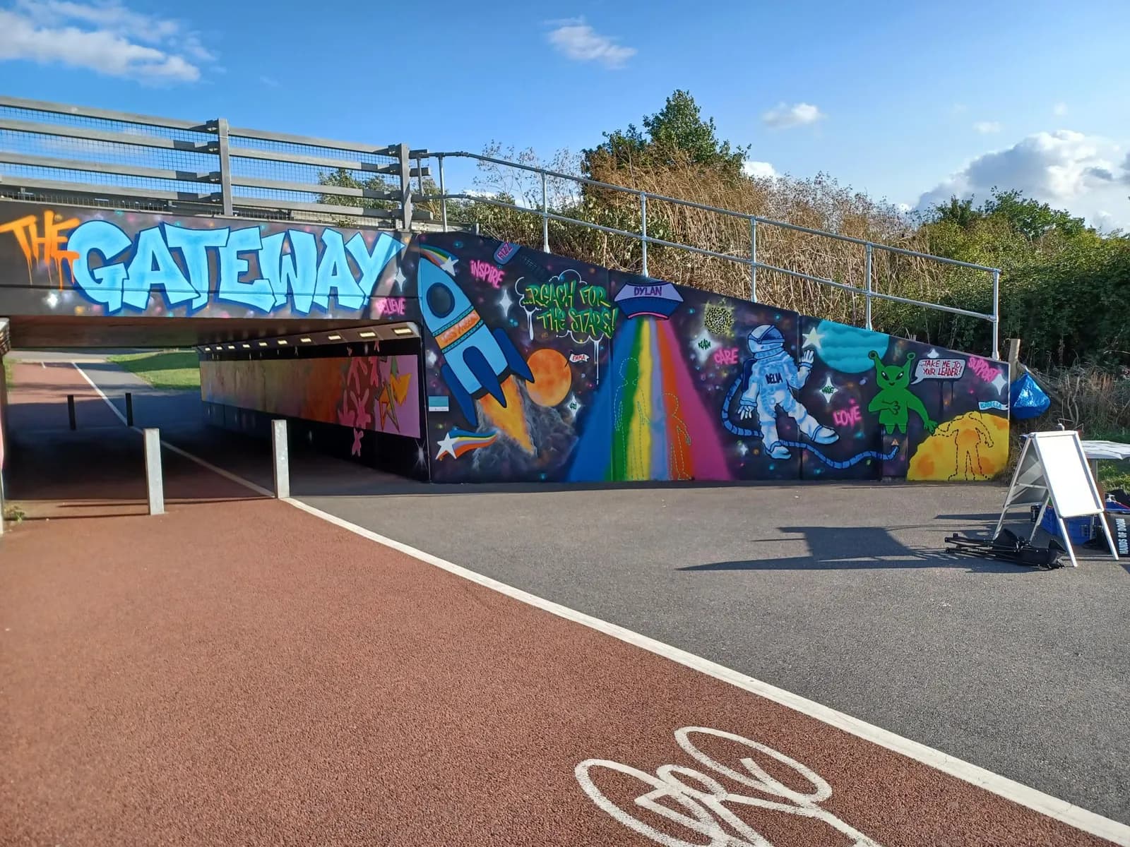 A vibrant underpass features a wall-length mural of a rocket, astronaut, and alien, along with the blue graffiti text "THE GATEWAY" above the entrance.