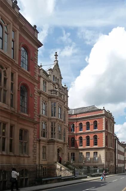 A grand, ornate sandstone building with intricate gothic detailing stands next to a simpler red brick building with arched windows along a city street under a partly cloudy sky.