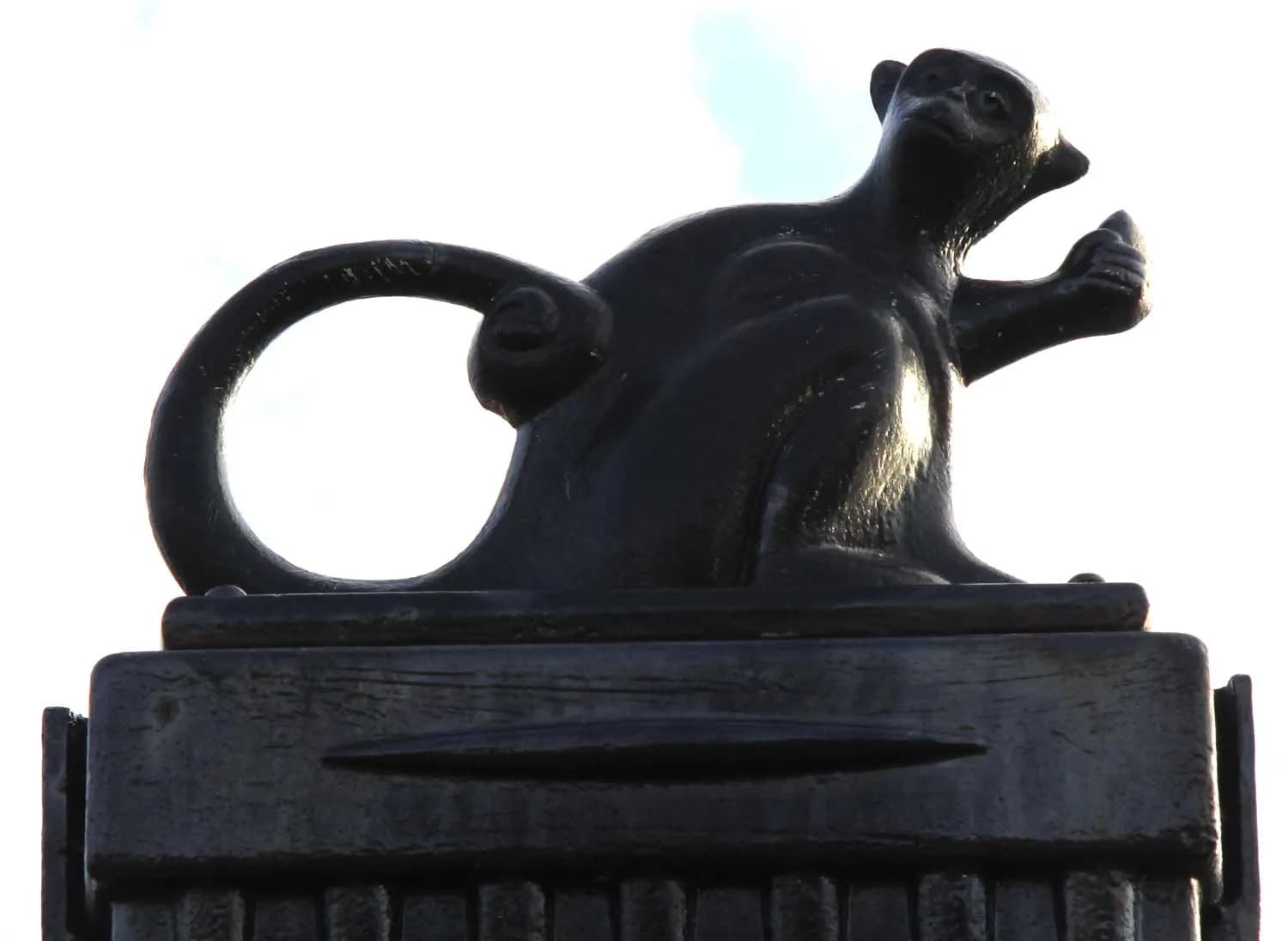 A dark monkey sculpture with its tail curled over its back gazes upwards, set against a bright, clear sky.