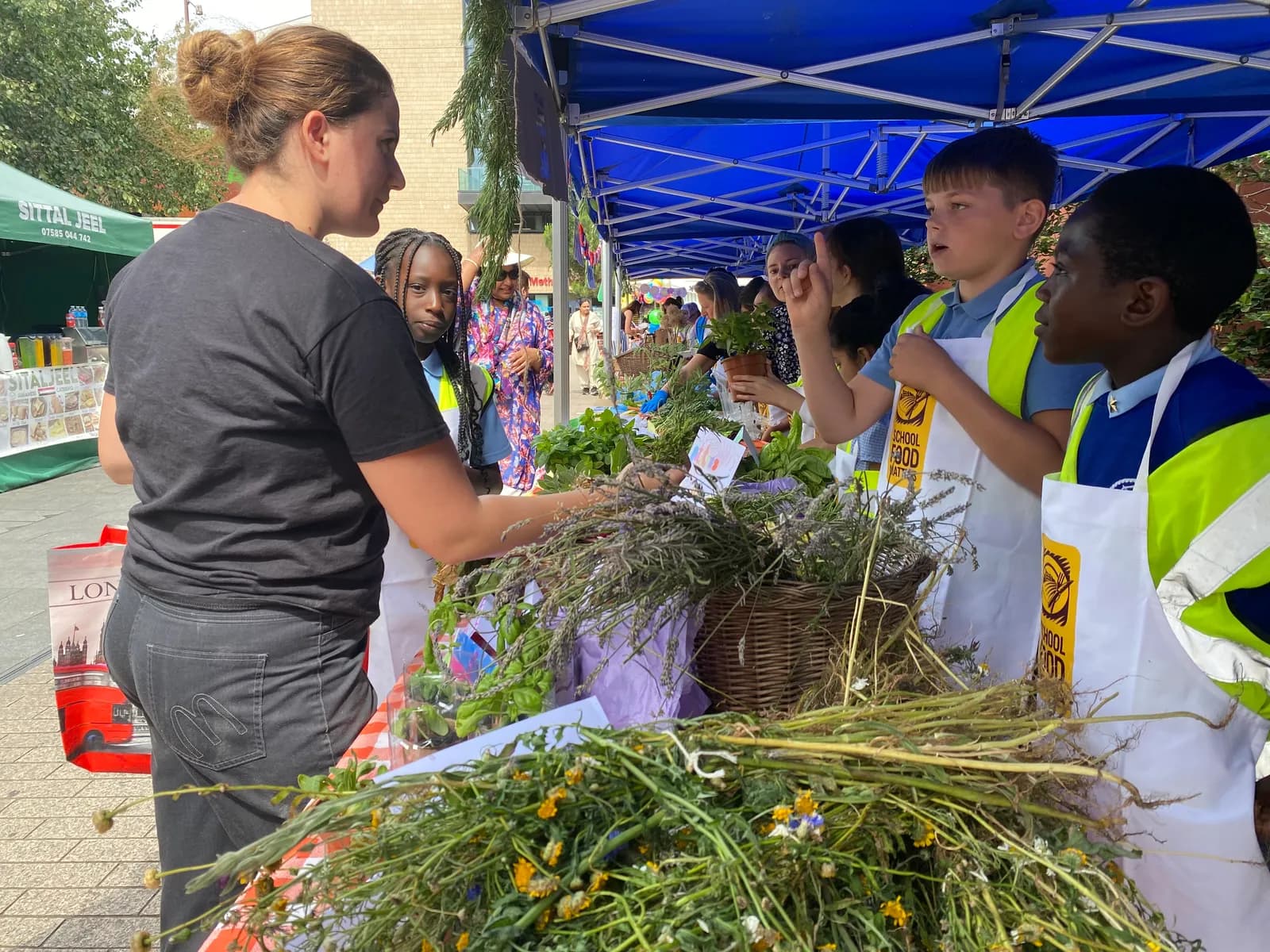 A woman speaks to school children under a blue tent, as they stand at a table covered in various plants and flowers.