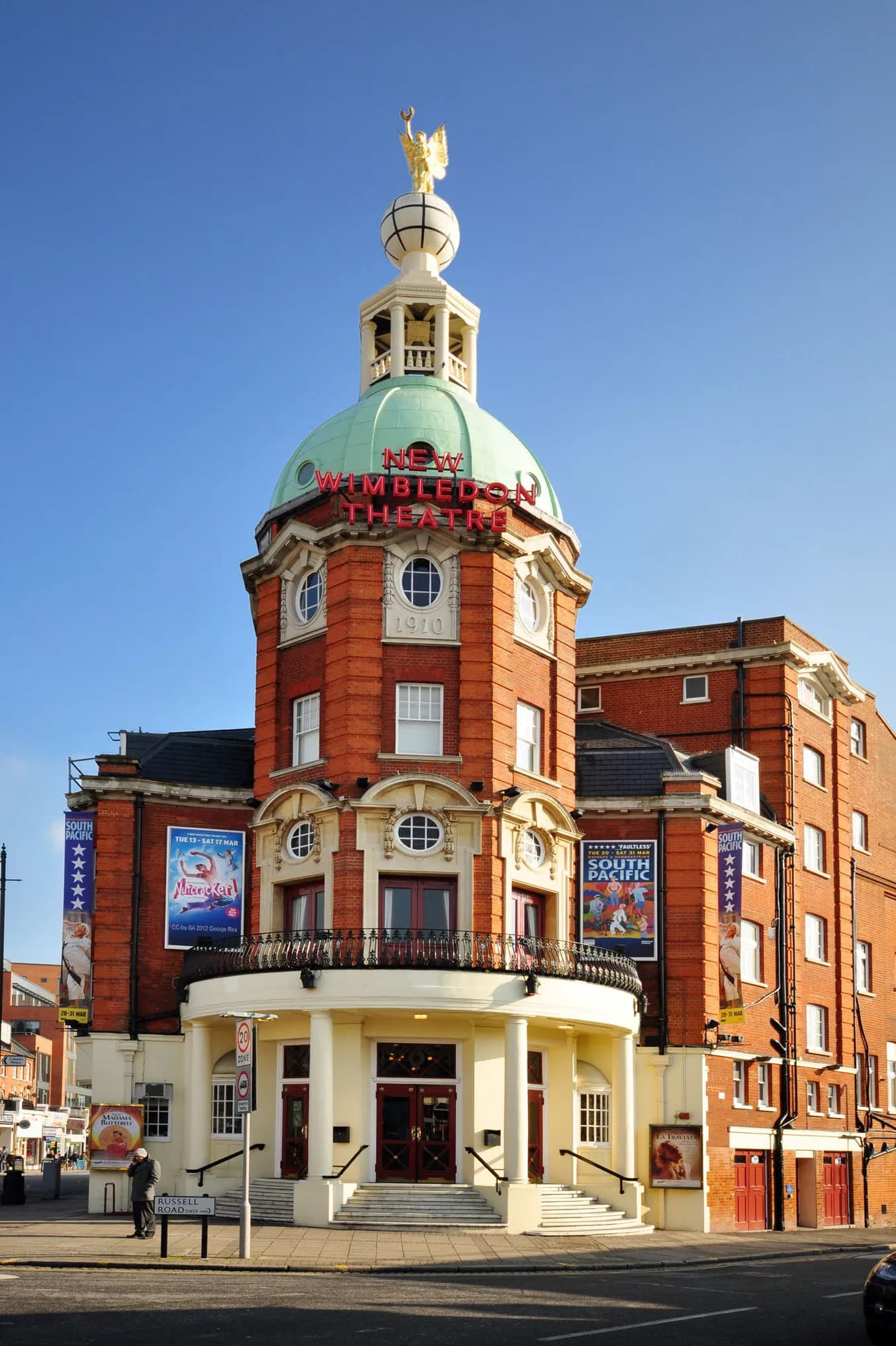 A grand, red-brick theater building with a prominent green dome topped by a golden statue under a clear blue sky.