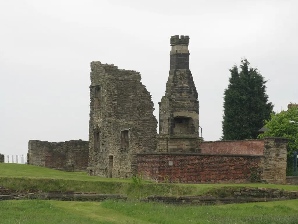 Remains of an old stone building with a prominent chimney stack standing on a grassy slope under a cloudy sky.