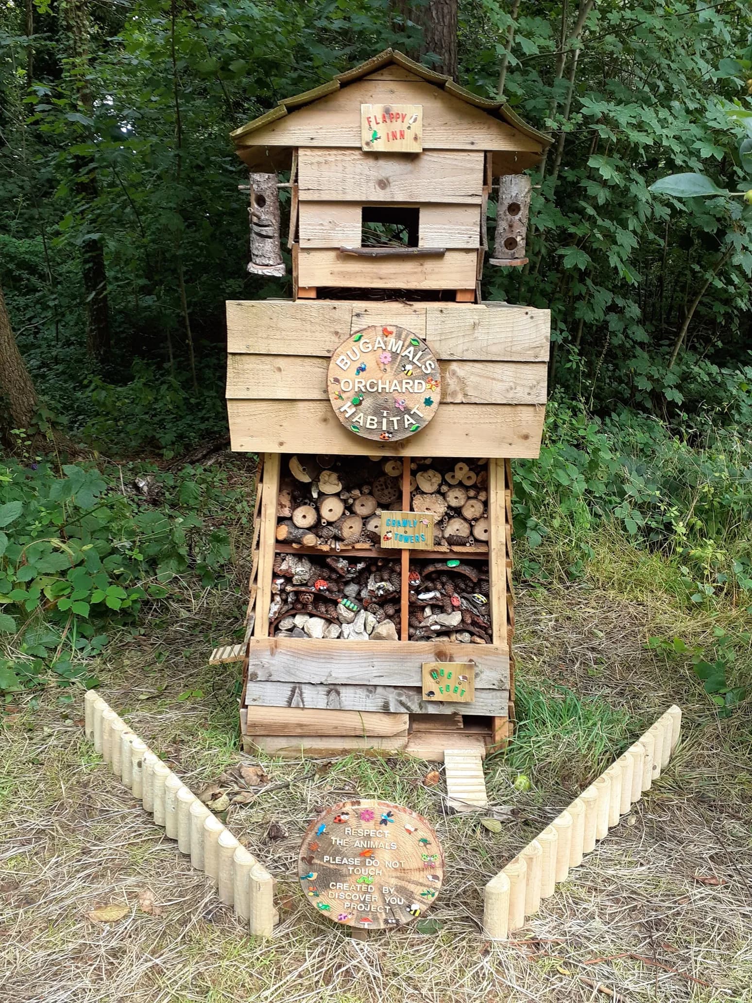 A tall homemade bug hotel made of wooden planks, filled with natural materials like logs and pinecones, stands in a grassy area surrounded by trees.