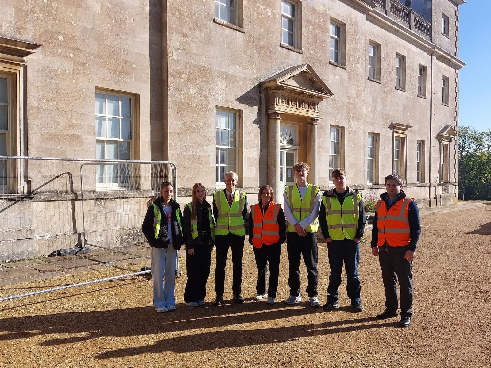 Seven people wearing safety vests stand in front of a grand stone building with many windows.