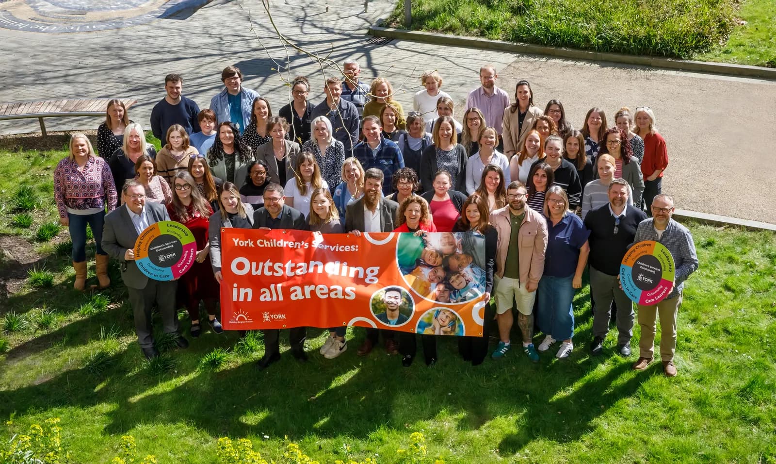 A large group of smiling adults, mostly women, stand on a grassy slope holding a banner that reads "York Children's Services: Outstanding in all areas."