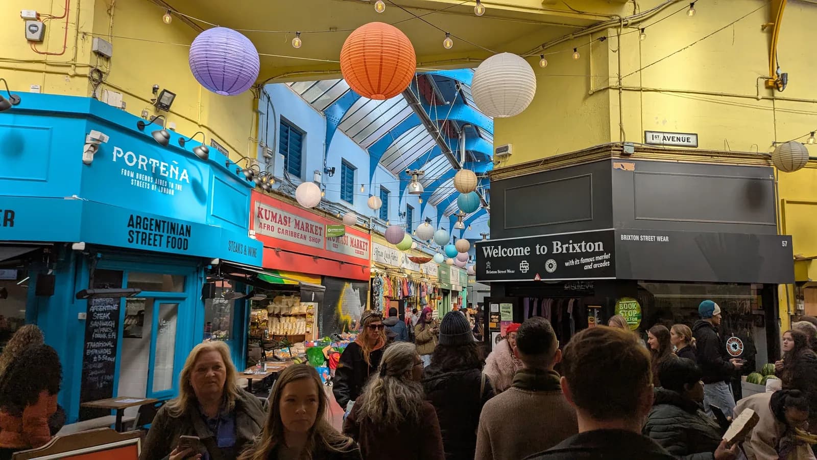 A busy indoor market filled with shoppers, food stalls, and various goods, decorated with colorful paper lanterns hanging from the ceiling.