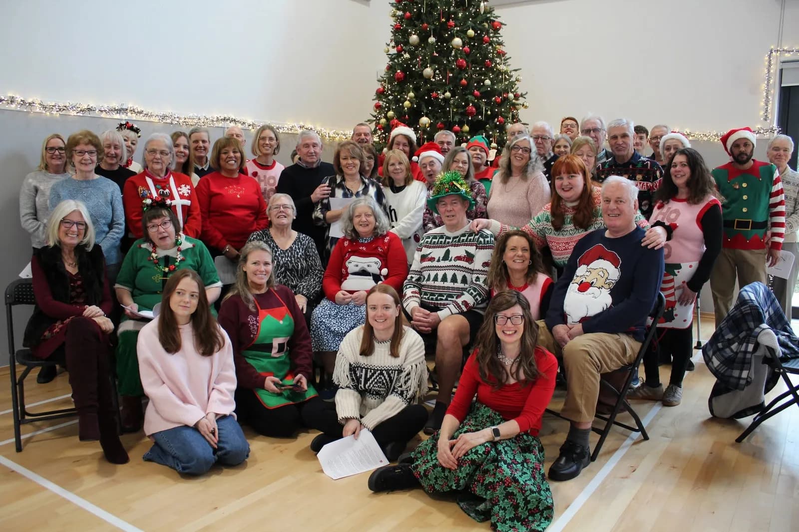 A large group of smiling adults wearing various festive Christmas sweaters and outfits, posing together in front of a brightly lit Christmas tree.
