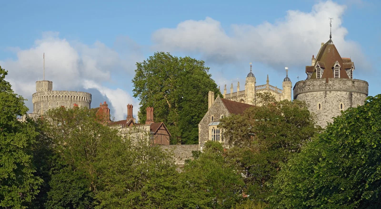 A view of Windsor Castle's stone towers and buildings partially obscured by lush green trees under a bright blue sky with white clouds.