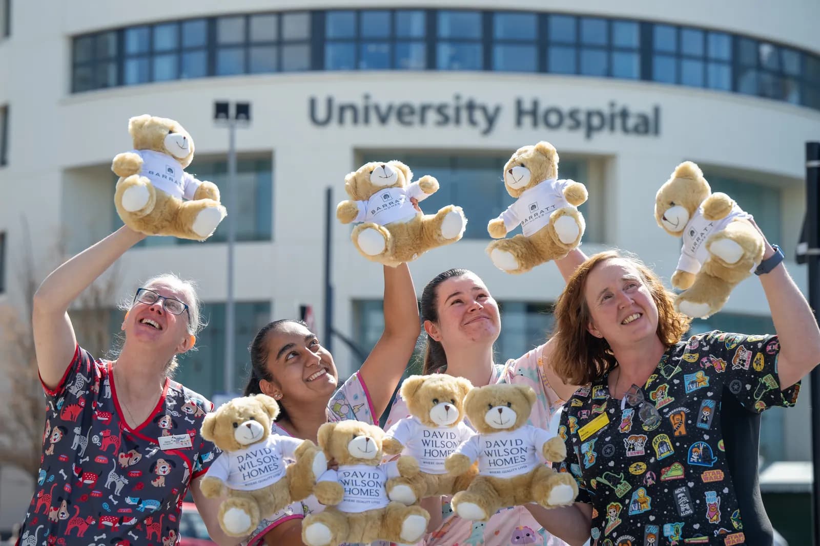 Several women in medical scrubs are holding up teddy bears, with some bearing the name "Barratt" and others "David Wilson Homes", in front of a building labeled "University Hospital".