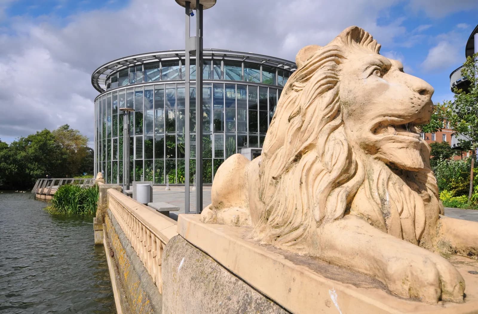 A majestic stone lion statue sits on a balustrade, facing right, with a modern glass botanical conservatory and a tranquil body of water in the background under a blue sky.