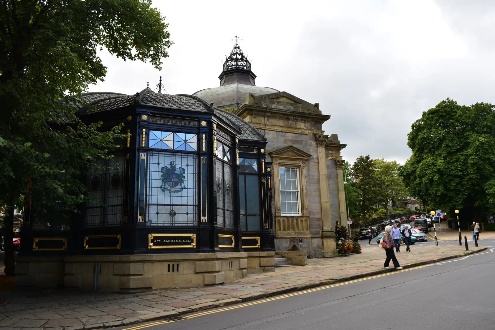 The black and gold facade of the Royal Pump Room Museum, featuring stained glass windows and an ornate dome, stands beside a road with a few pedestrians and green trees under an overcast sky.