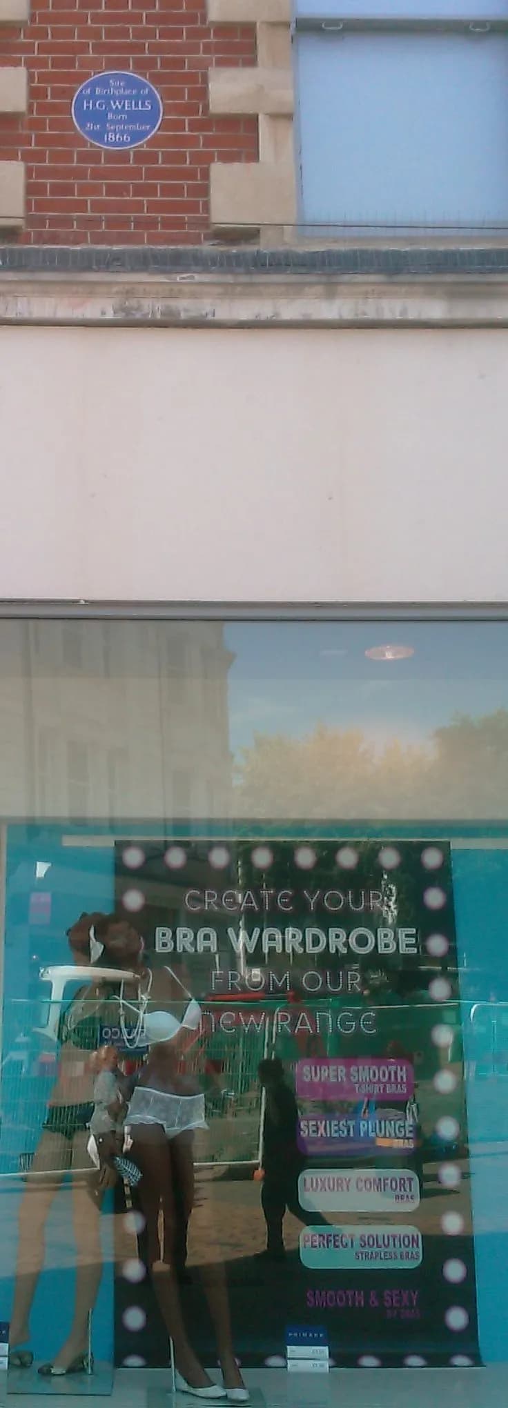 A blue plaque on a brick building marks the birthplace of H.G. Wells, with a window display below advertising 'Create Your Bra Wardrobe'.