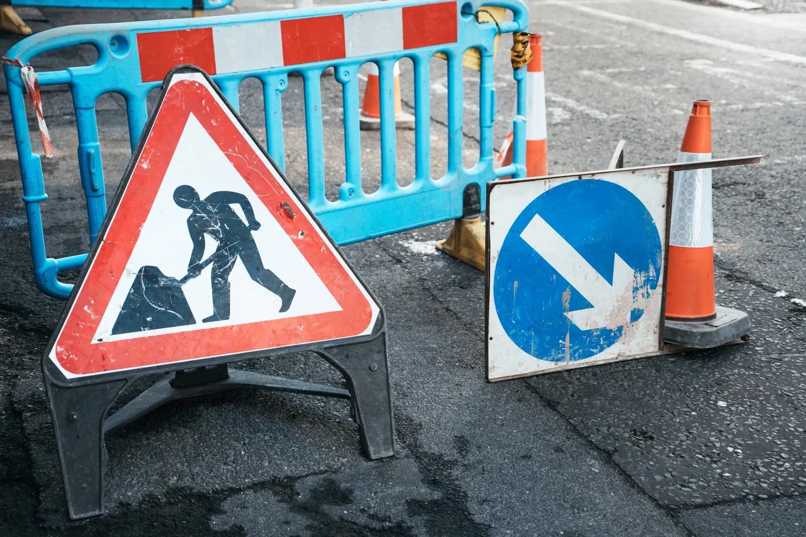 A red and white triangular road sign with a black silhouette of a person digging, next to a blue and white circular sign with a white arrow pointing downwards and to the right, on an asphalt road with a blue barrier and orange cones in the background.
