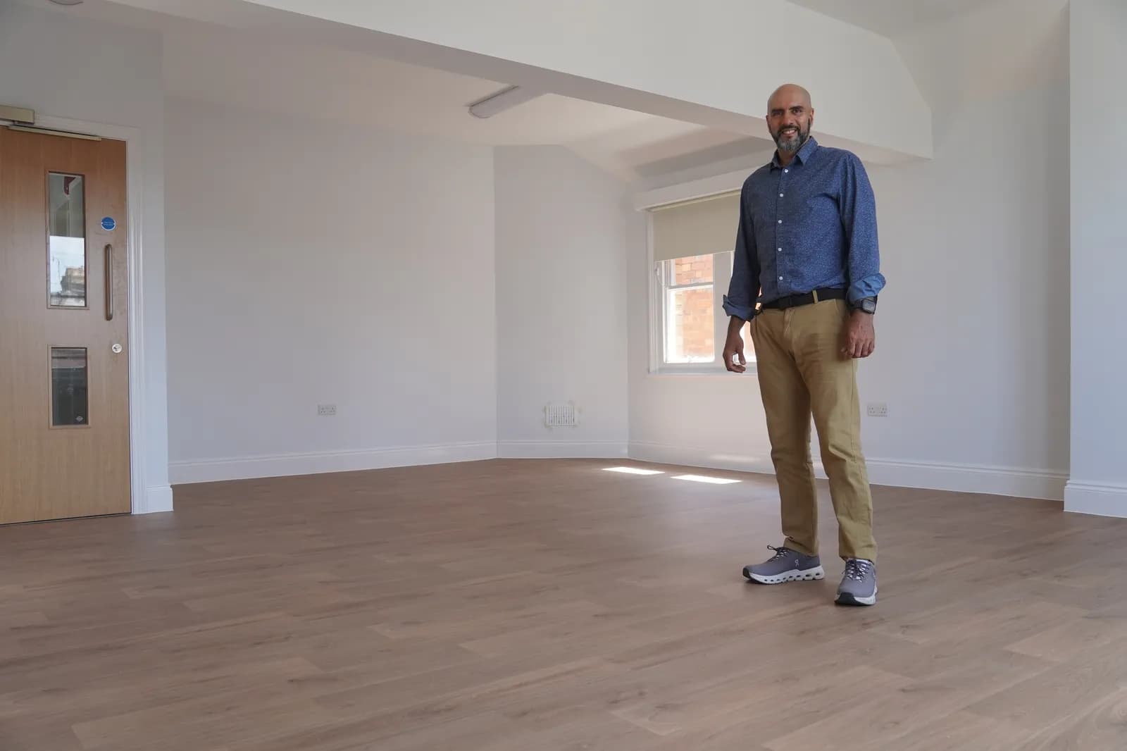 A man with a beard and dark hair stands in a large, empty, white room with wooden floors. He is wearing a blue button-up shirt, tan pants, and grey sneakers. There is a window behind him and a wooden door with an emergency exit bar to his left.