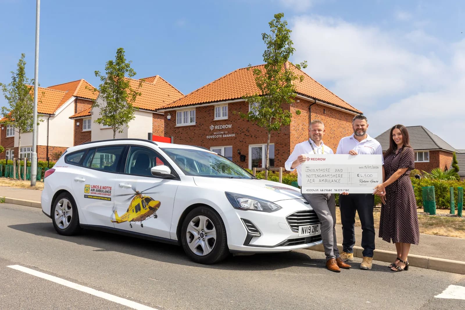 Three people holding a large check in front of a white car with a helicopter graphic and houses in the background.