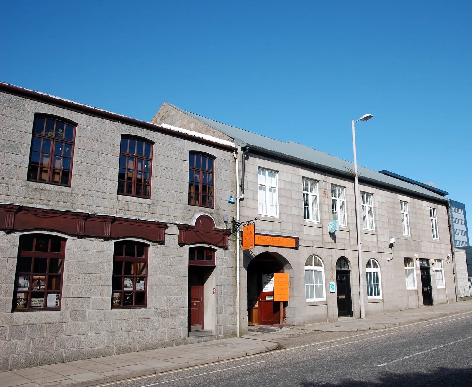 A long, two-story stone building with several windows and doorways sits next to a paved road under a clear blue sky.