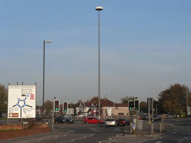 A complex road intersection, known as 'The Magic Roundabout', with multiple traffic lights, directional signs, and several cars navigating its lanes under a clear sky.