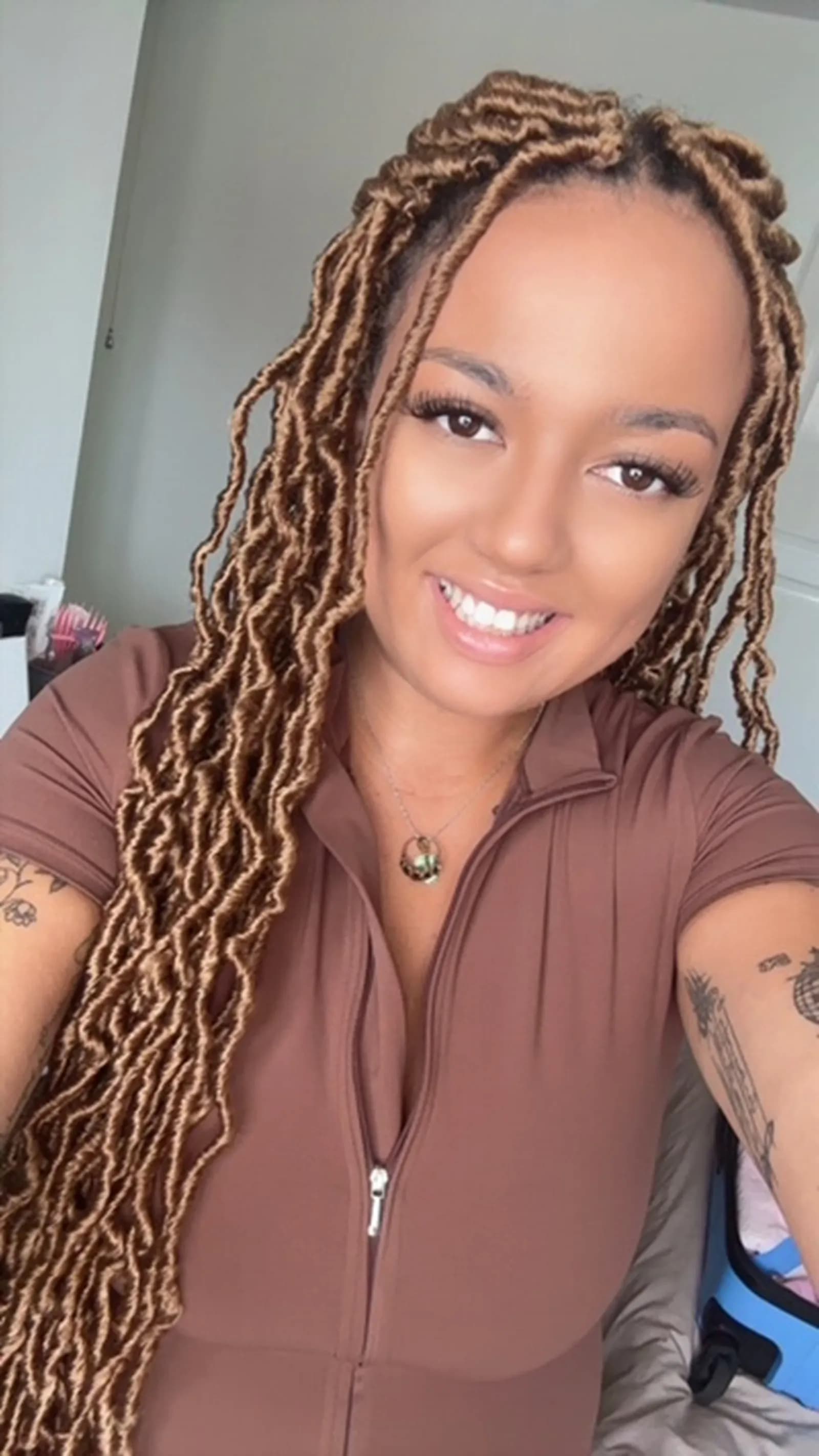 A smiling woman with long brown dreadlocks and tattoos on both arms, wearing a brown zippered top and a pendant necklace.