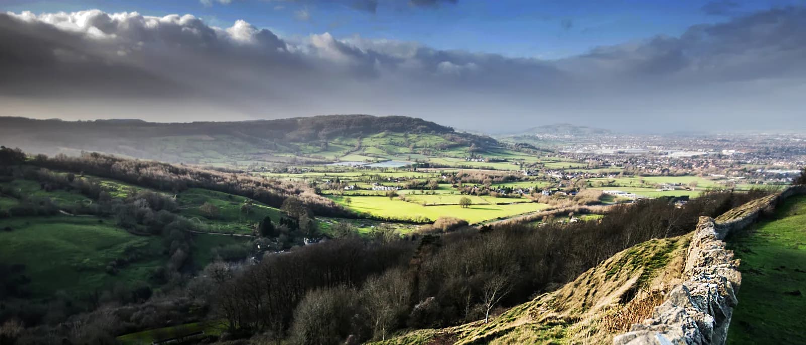 A panoramic landscape shows rolling green hills and fields in a valley under a partially clouded sky, with a town visible in the distance and a stone wall on a grassy slope in the foreground.