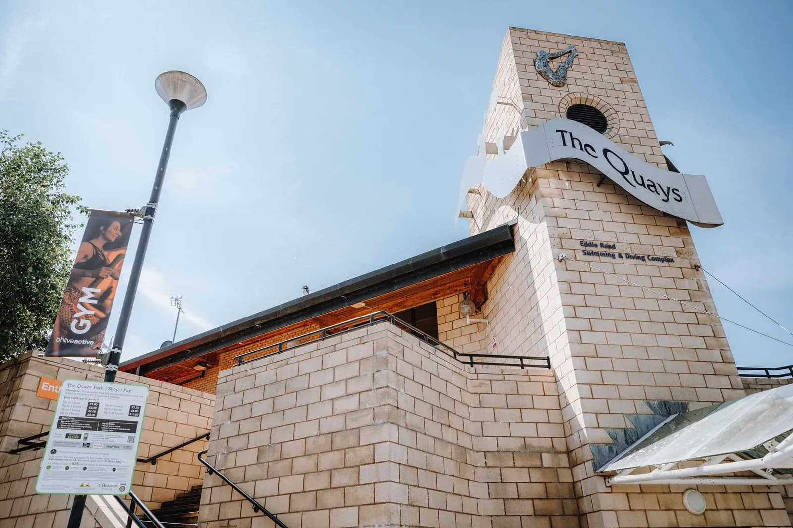 The entrance to The Quays swimming and diving complex, a building made of light-colored bricks with a large sign that says 'The Quays' and a banner for 'GYM' attached to a lamppost.
