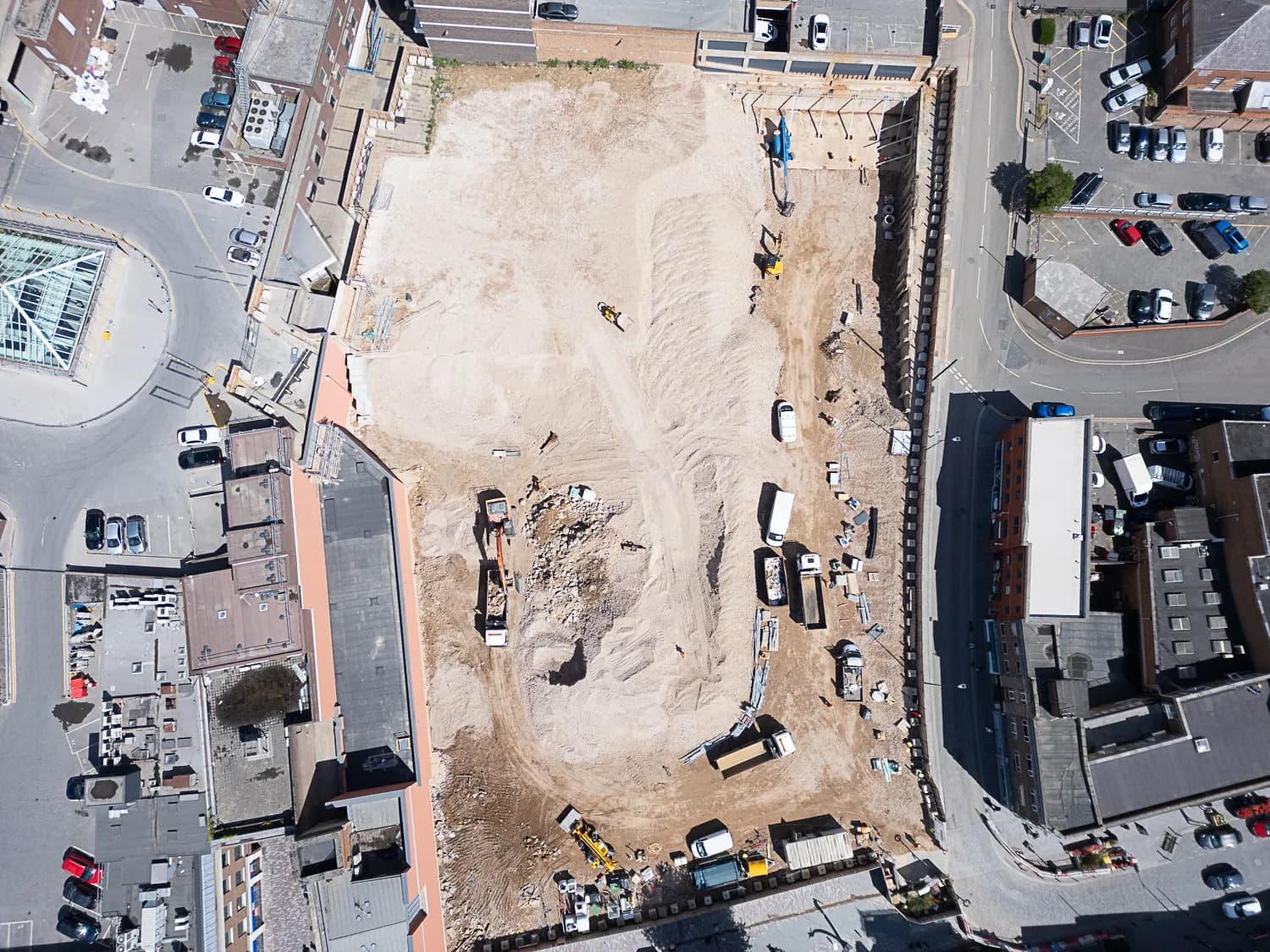 An aerial view of a large, cleared construction site with multiple construction vehicles, surrounded by roads, buildings, and parked cars.