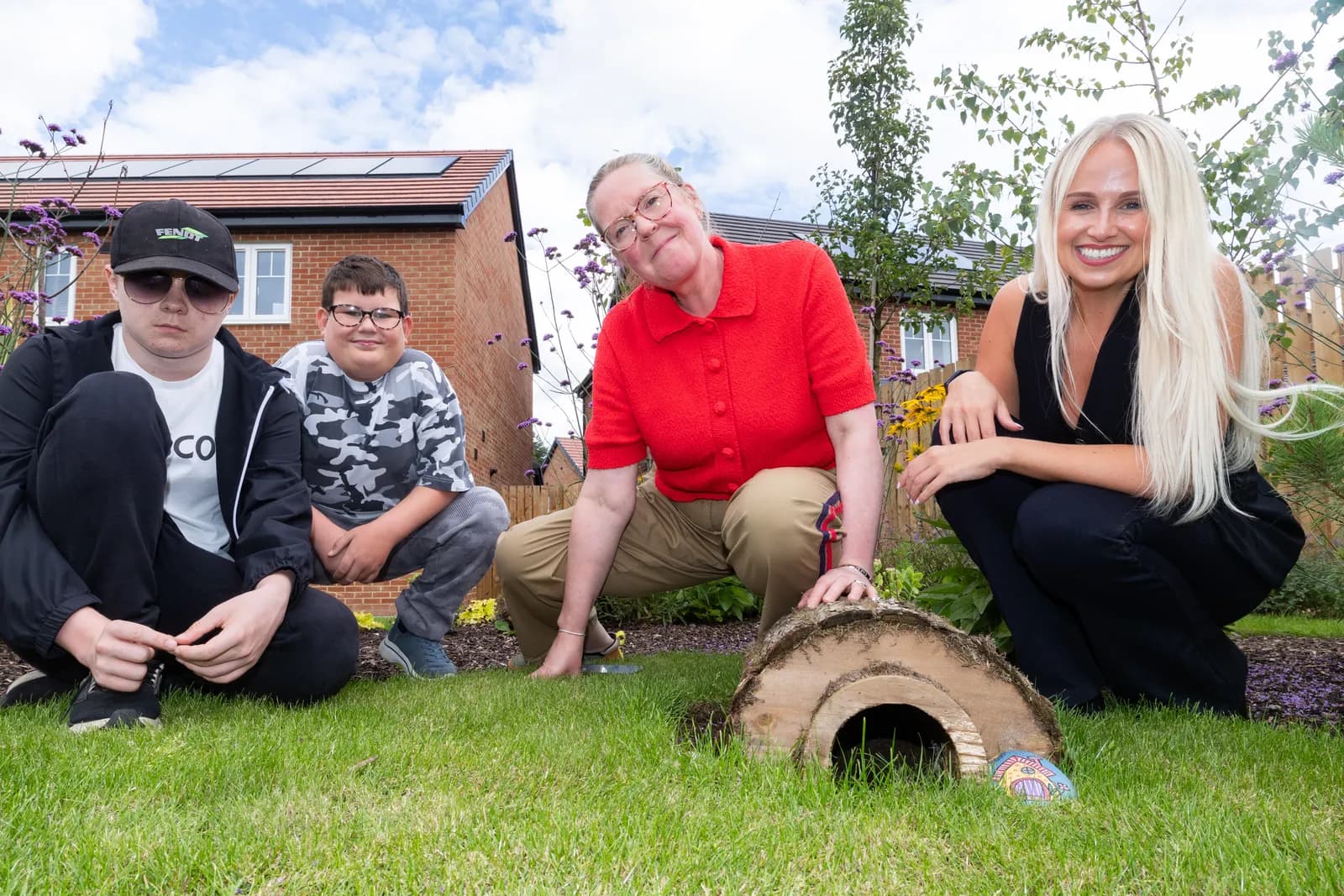 Three adults and one child posing on a grassy lawn with houses and greenery in the background, next to a small wooden hedgehog house.