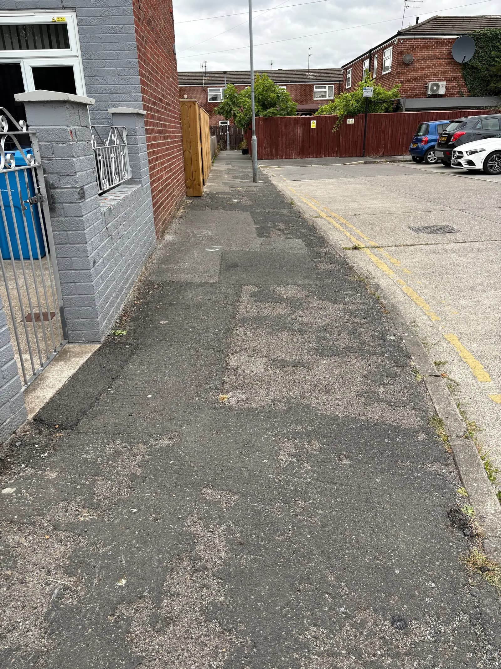 A long, narrow asphalt pathway next to a brick house and a red fence, leading towards a residential area with parked cars.