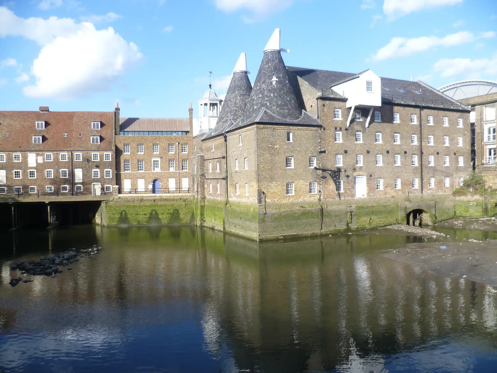 A historic brick building with two conical roofs stands at the edge of a canal, reflecting in the water, with other buildings and a clear sky with scattered clouds in the background.