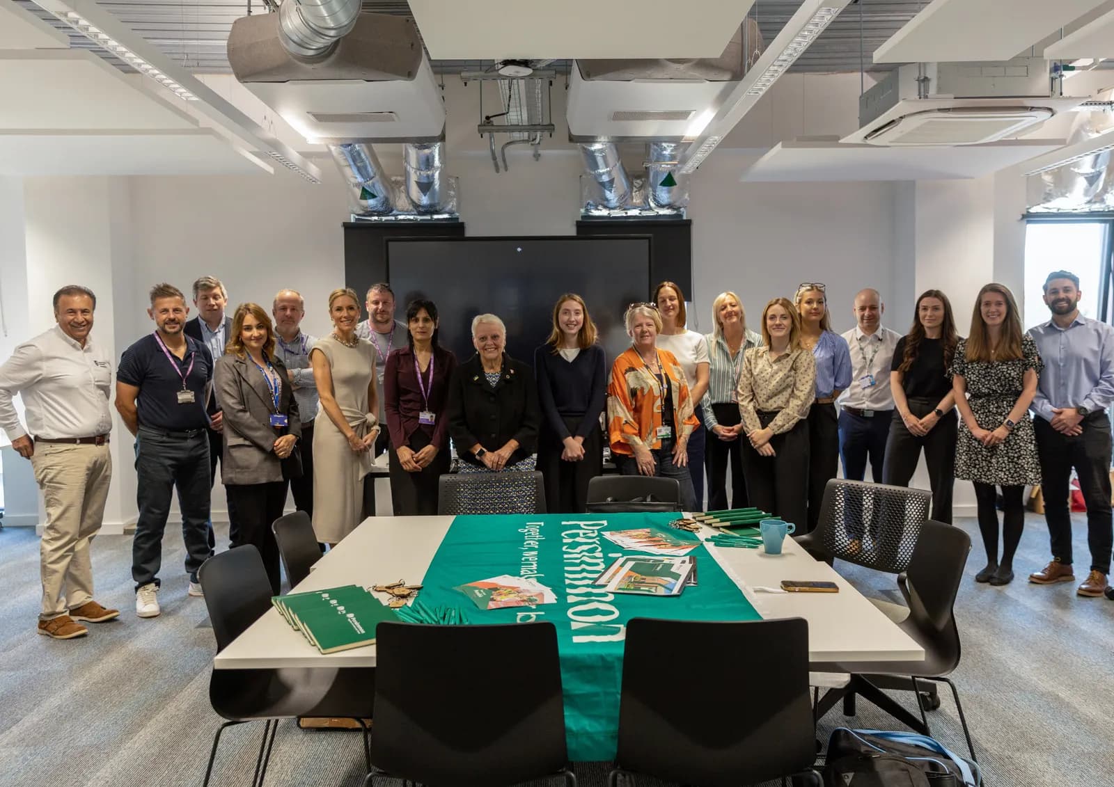 A group of diverse people stand in a modern office, some wearing lanyards, with a table in the foreground covered by a green banner.