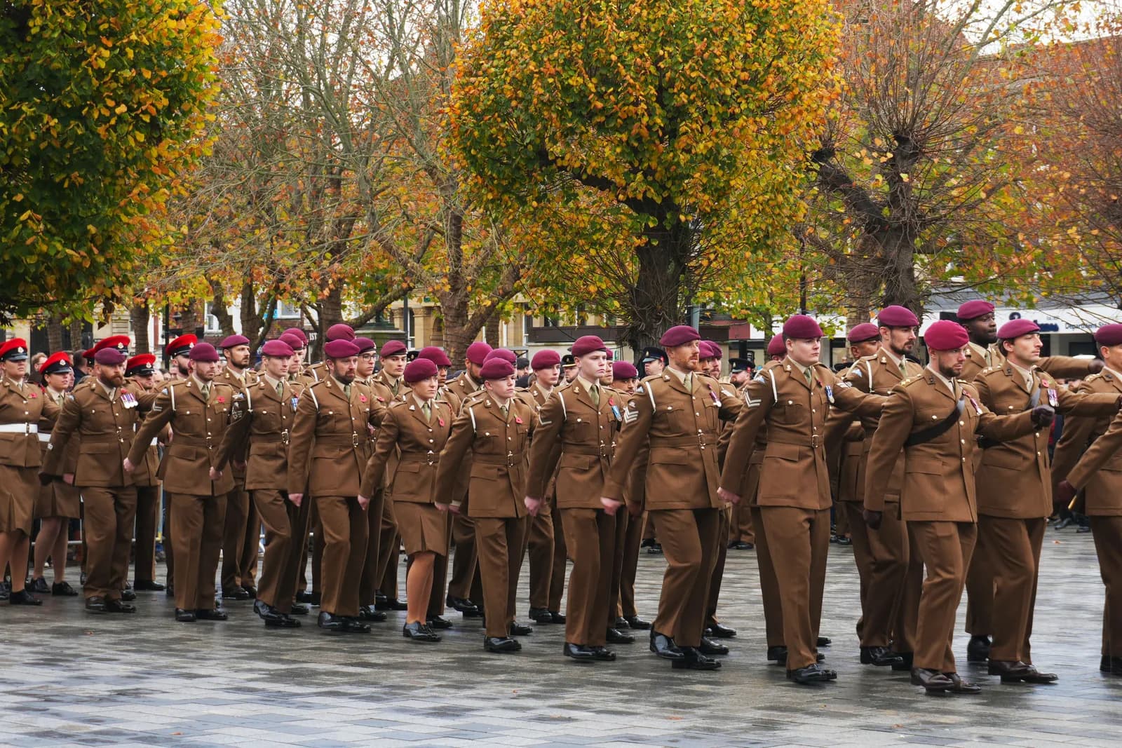 A large group of soldiers in brown uniforms and red berets march in formation on a paved area with trees featuring autumn foliage in the background.