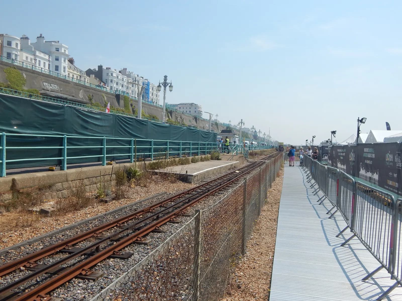 A long shot of an empty train track running alongside a fenced walkway, with white buildings visible on a hill in the background.
