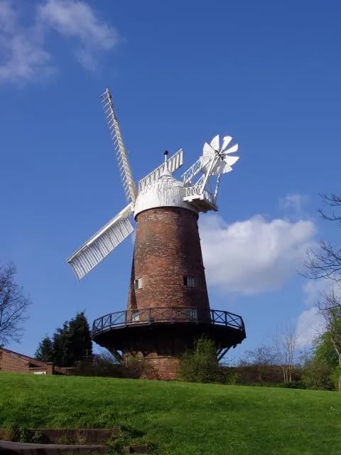 A historic brick windmill with white sails stands tall on a grassy hill against a clear blue sky with some white clouds.