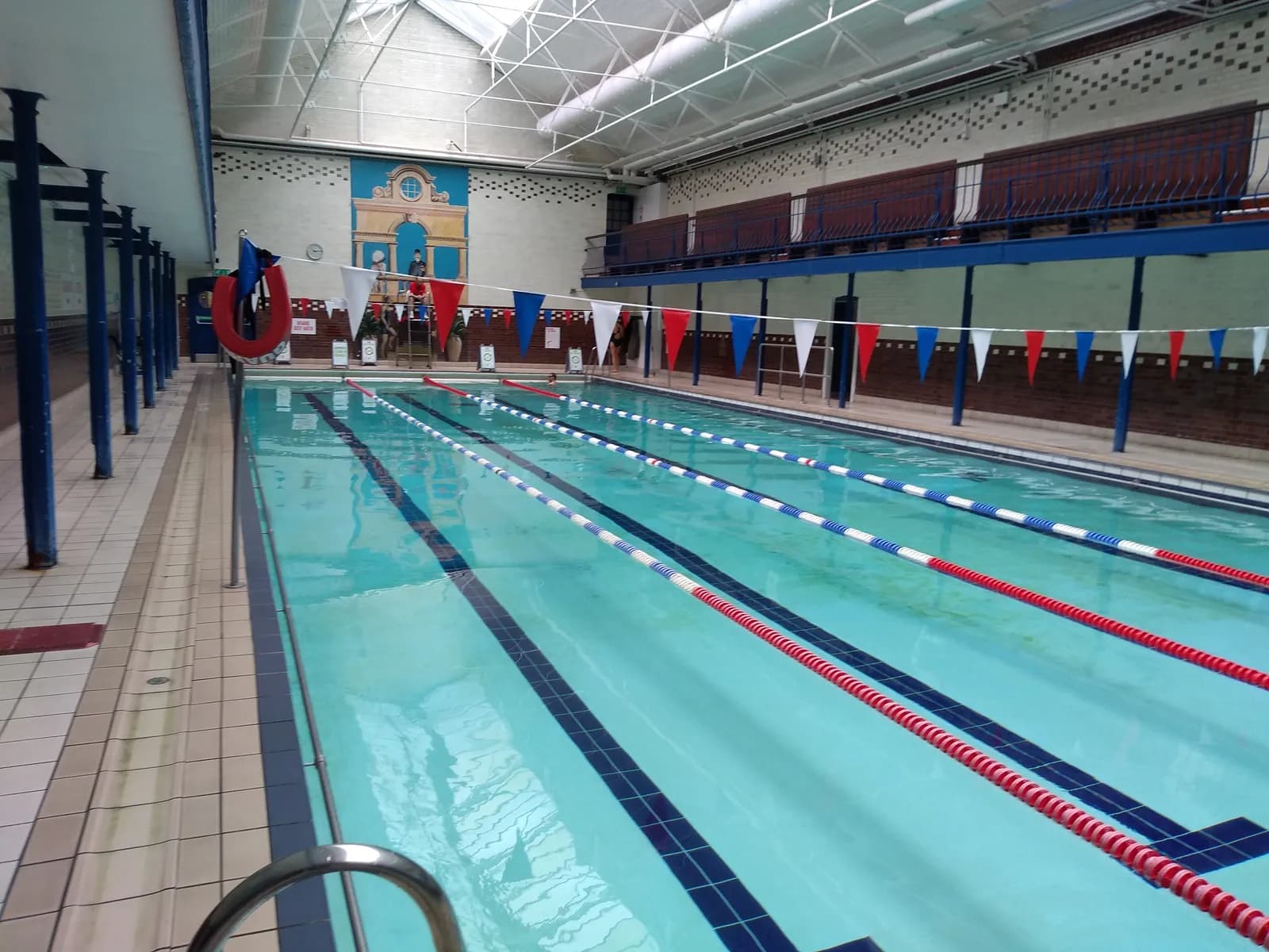 An empty indoor swimming pool with red, white, and blue lane lines and a colorful mural on the far wall.