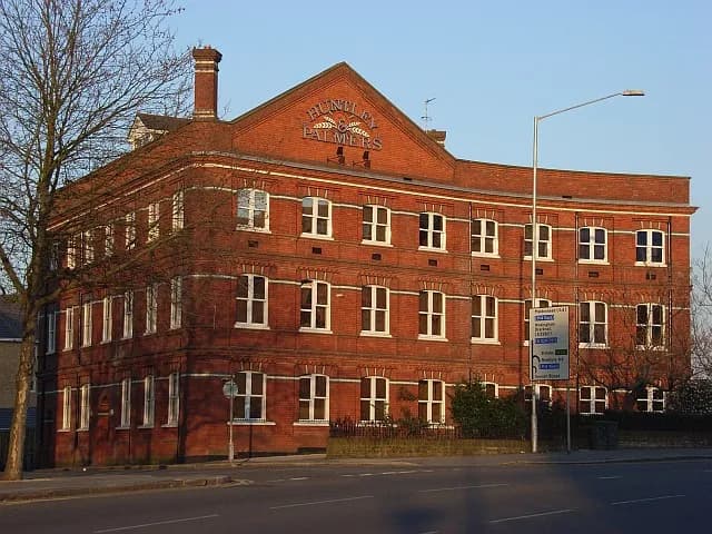A large red brick building with many windows and an arched sign on its facade, with a street and bare trees in the foreground, under a clear sky.