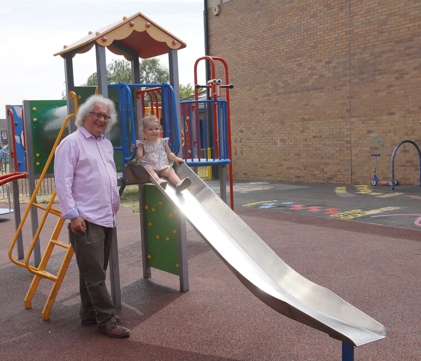 A man and a toddler stand on a brightly colored playground with a slide and climbing structures, with a brick building in the background.
