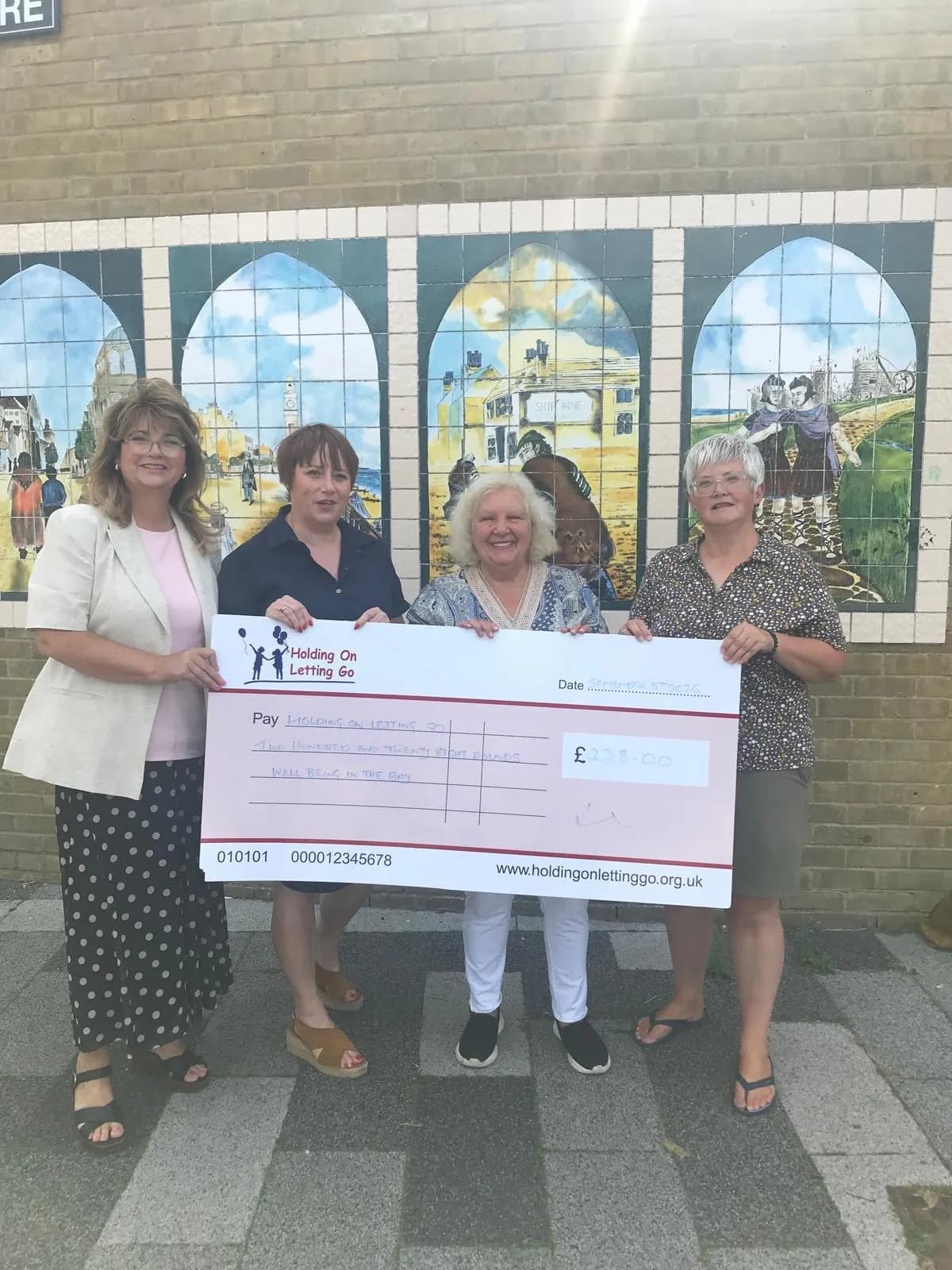 Four women hold a large check for 233 pounds in front of a brick wall with a painted mural.