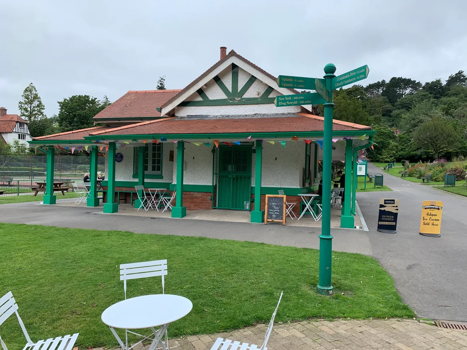 A small white building with green accents, a red-tiled roof, and a covered porch with green pillars, decorated with colorful triangular flags, sits next to a path with a green signpost showing directions, all surrounded by green grass and trees under a cloudy sky.
