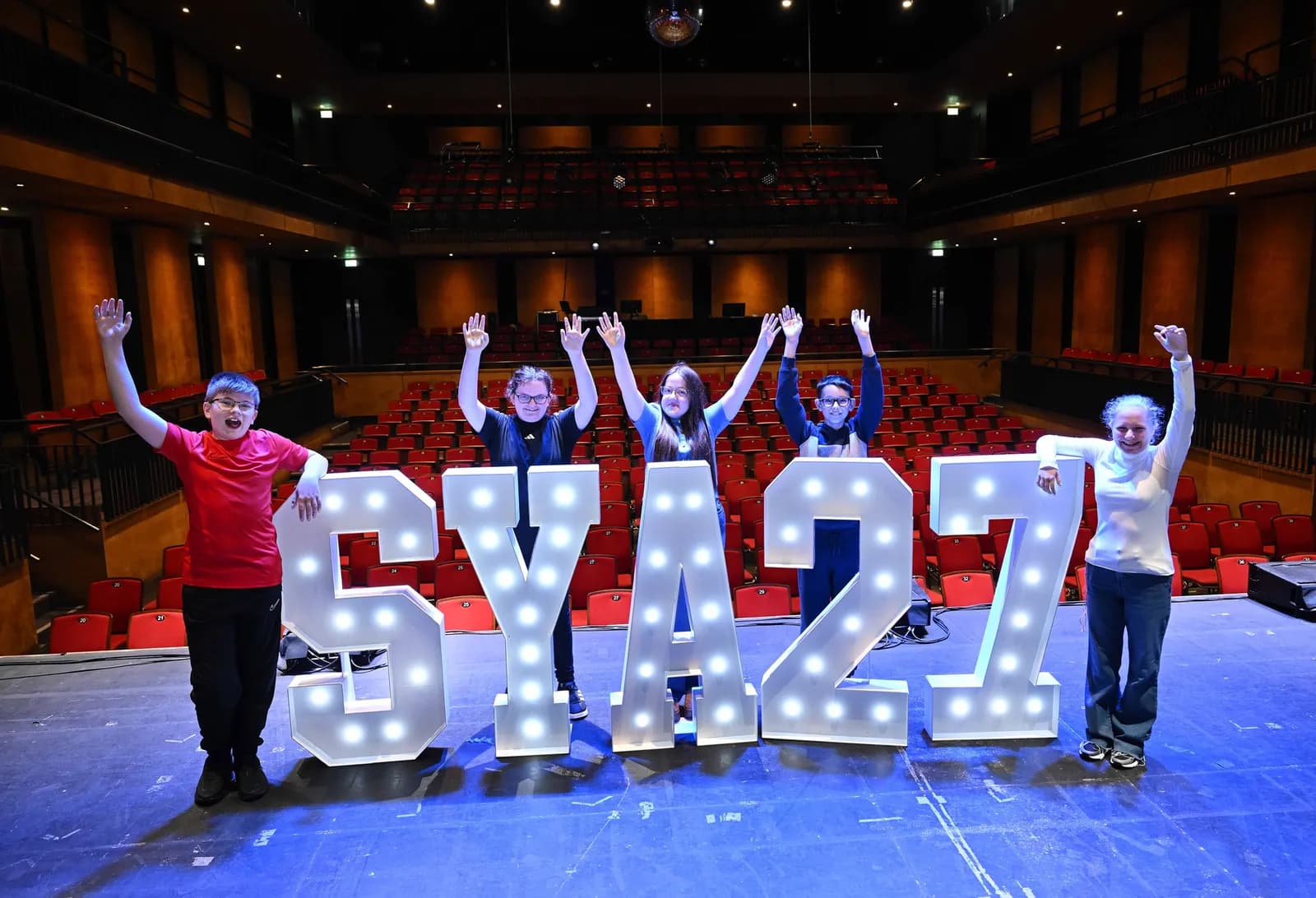 Five children standing on a stage with large, illuminated letters spelling "SYAZ 27" in front of them, with an empty theater audience visible in the background.