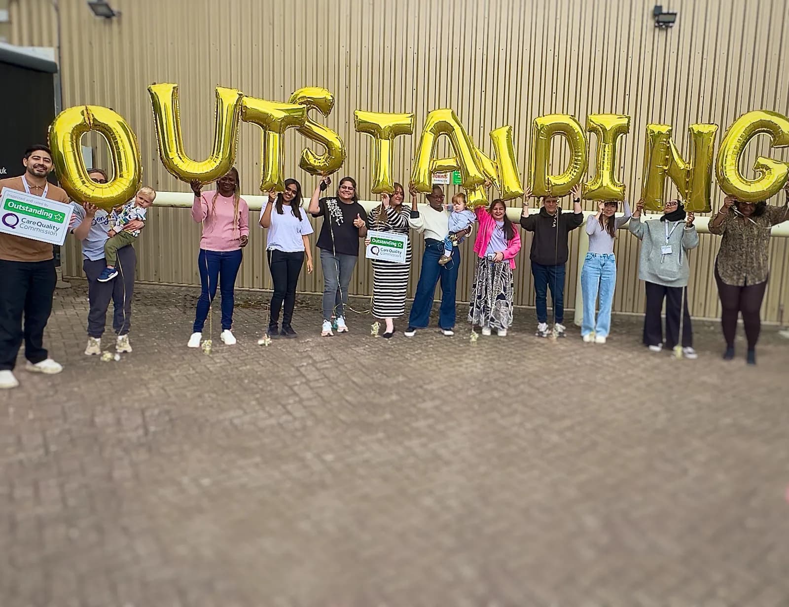 A diverse group of people, including adults and children, holding up golden balloons that spell out the word "OUTSTANDING" in front of a beige corrugated wall.